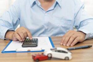 Car Insurance Concept: A man uses a calculator on a wooden table to assess damage costs from a car accident, with a model car illustrating the accident. 