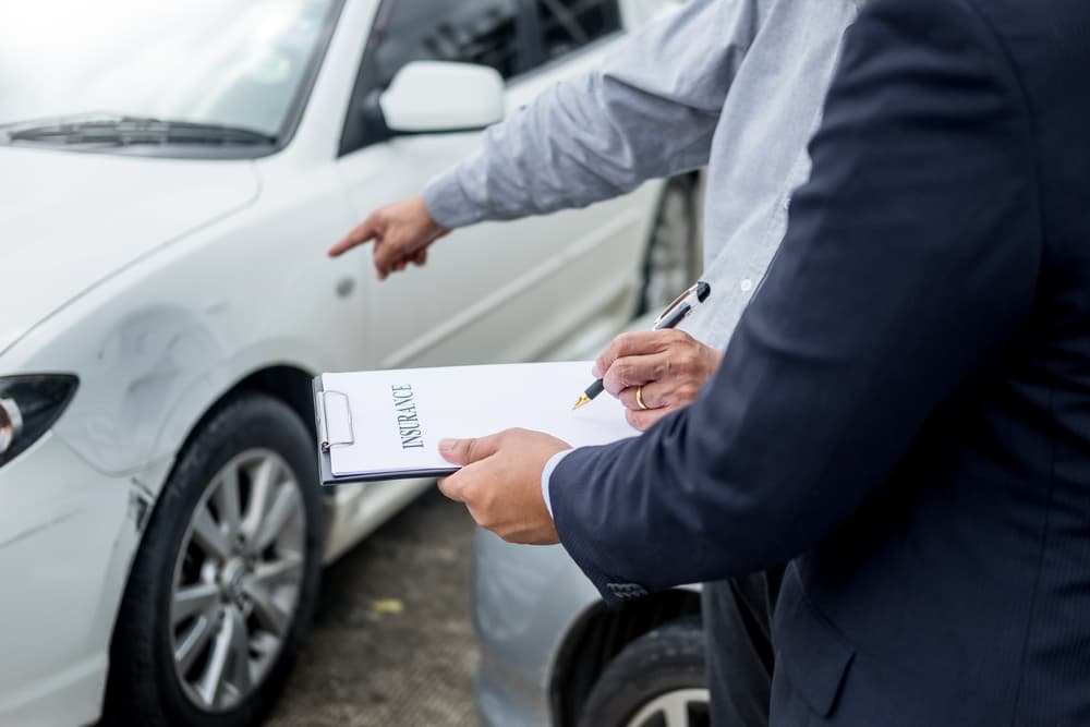 An insurance agent records details on a clipboard while inspecting a car involved in an accident, assessing and processing the claim.