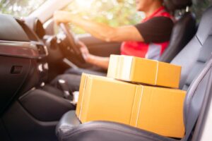 A delivery driver is driving a van with parcel boxes on the seat, preparing to deliver packages to customers outside the warehouse.