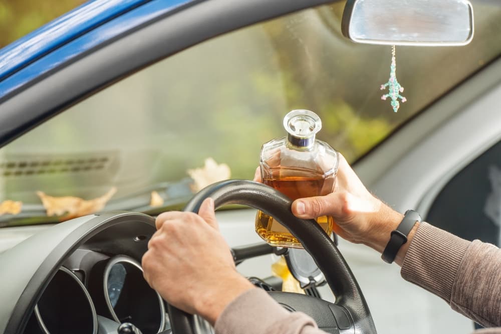 The driver sits behind the wheel, holding an alcoholic beverage, breaking the law by drinking while driving.