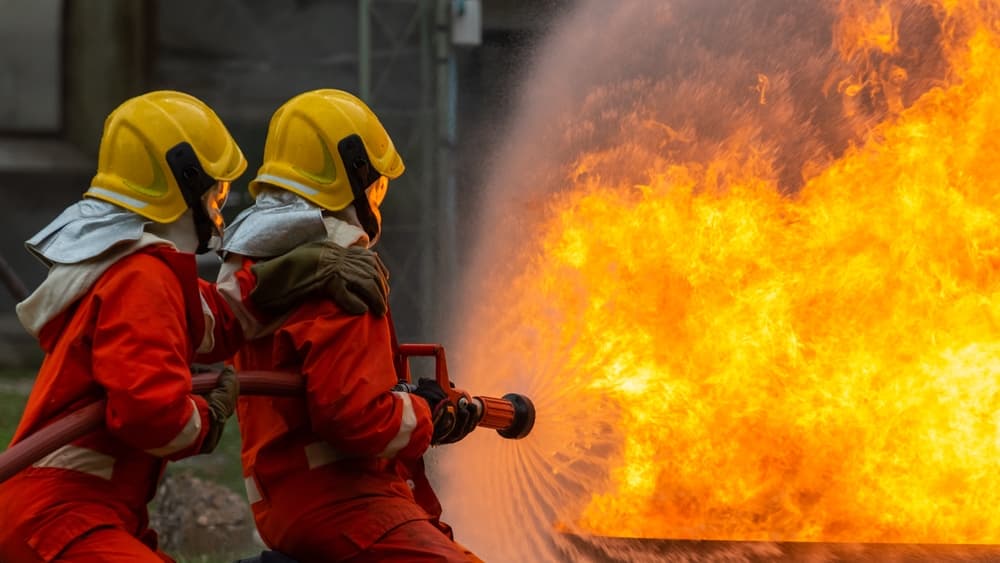 Firefighters in full gear work together on a rescue mission, using a hose to extinguish intense flames within a burning building.