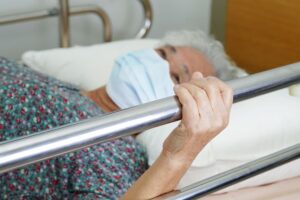 An elderly Asian woman lies in a hospital bed, holding the bed rail with a hopeful expression as she waits for her family to visit.