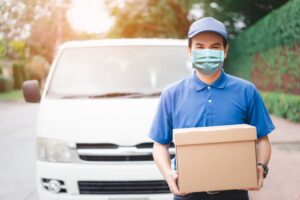 A postal delivery courier wearing a protective face mask stands in front of a cargo van, holding a package for delivery during the COVID-19 outbreak.